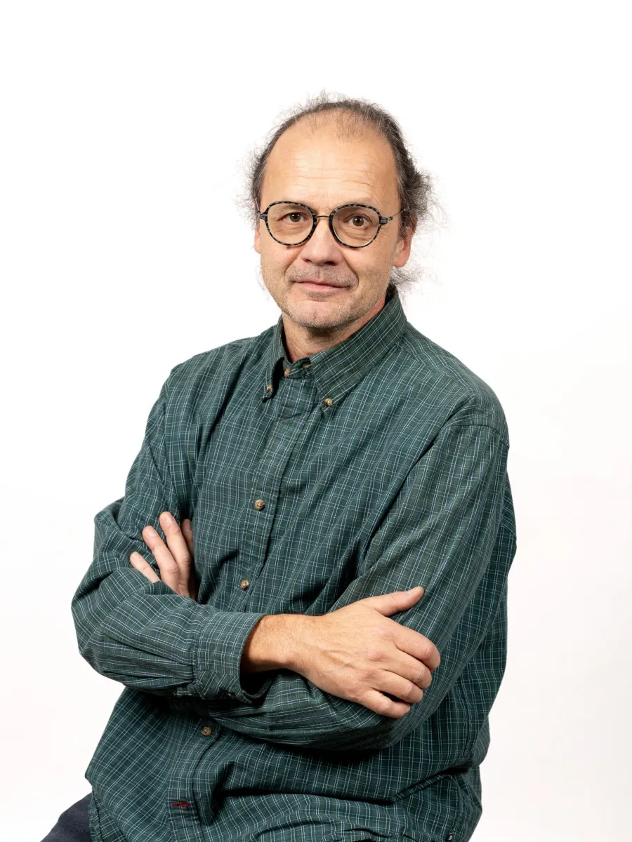 Alex Fekete - A man with glasses is wearing a green plaid shirt and jeans. He is sitting with arms crossed against a white background.