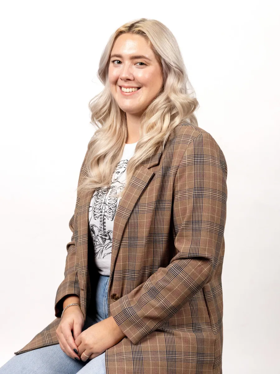 Alison Taras - A person with long blonde hair sits on a stool against a white background. They are wearing a plaid blazer over a graphic t-shirt and light blue jeans, and are smiling towards the camera.