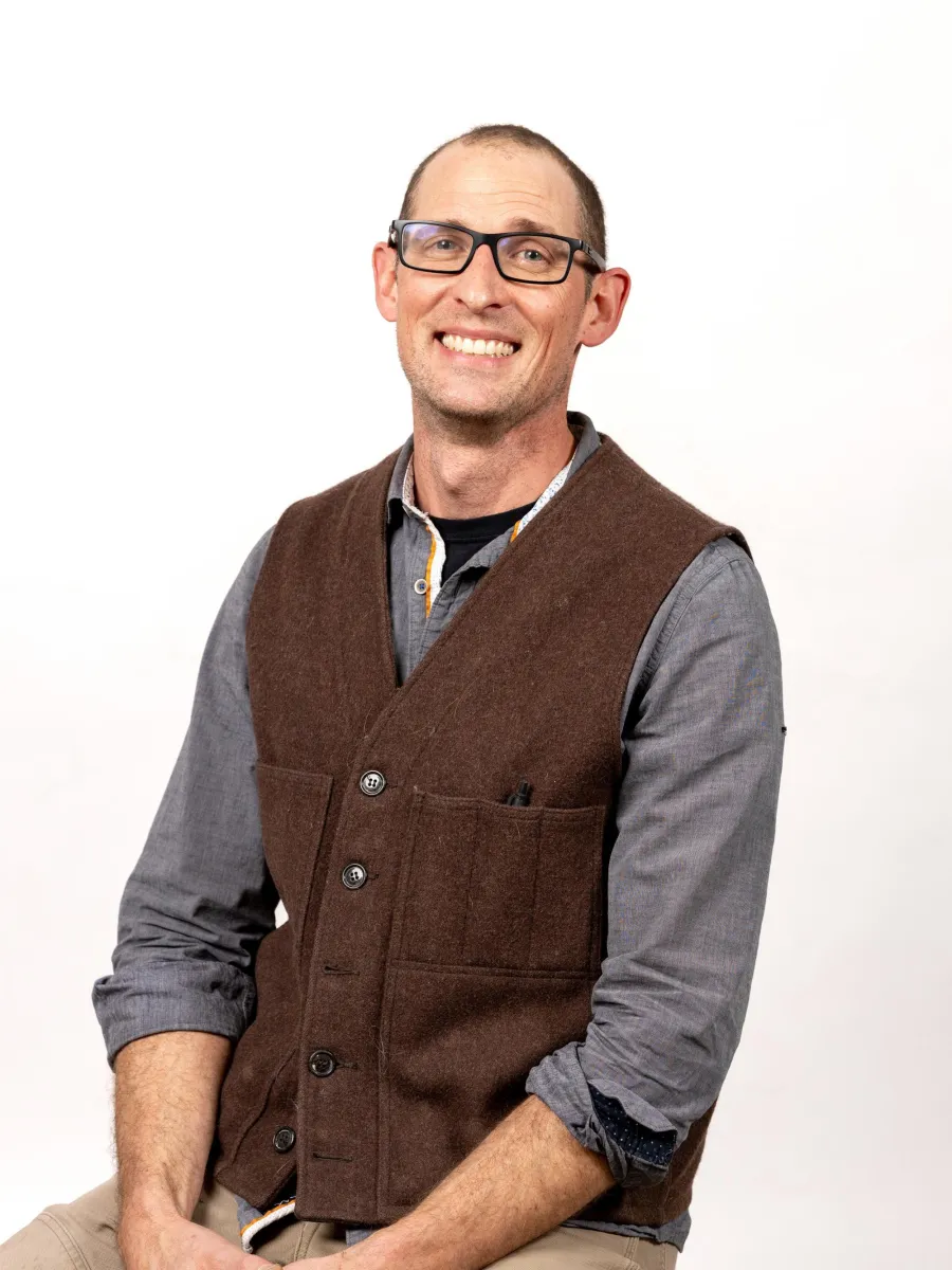 Brian Kakas - A man with glasses and short hair smiles while sitting. He is wearing a brown vest over a gray shirt and beige pants. The background is plain white.