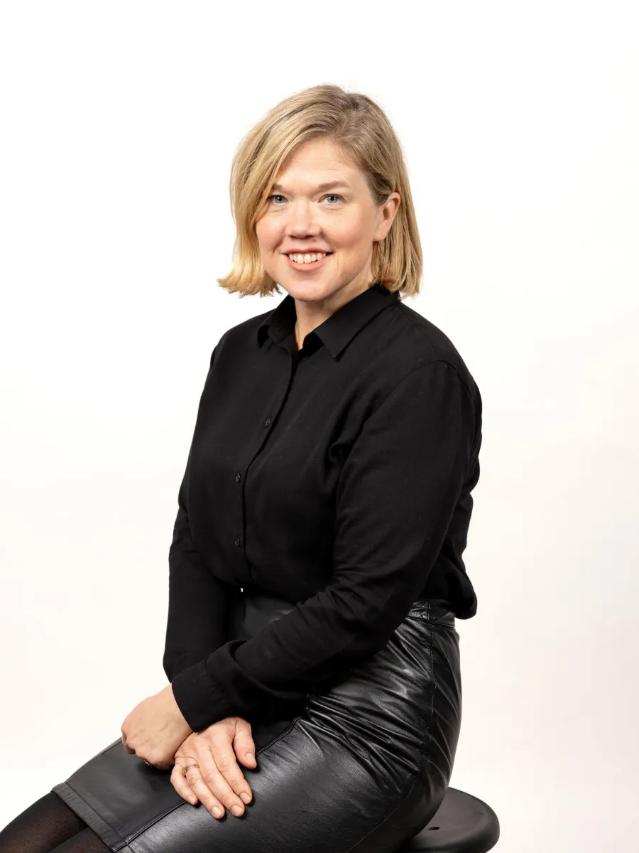 Emily Lanctot - A person with shoulder-length blonde hair is sitting on a stool. They are wearing a black long-sleeve shirt and a black leather skirt, smiling at the camera against a plain white background.