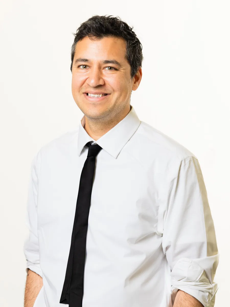 Ezra Asohan - Smiling person with dark hair wearing a white shirt and black tie, standing against a plain light background.