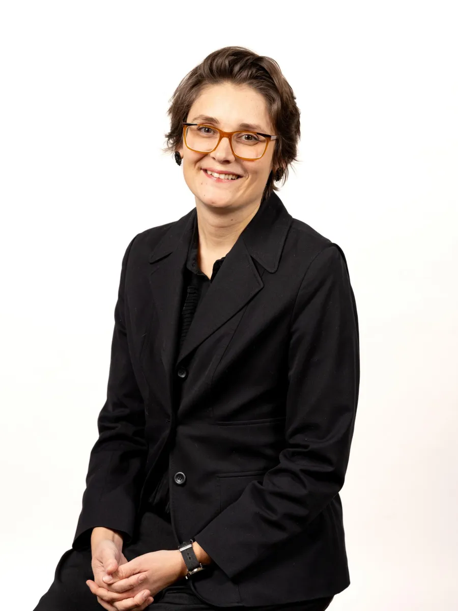 Gabrielle McNally - A person with short brown hair and glasses is smiling while seated against a plain white background. They are wearing a black suit and have their hands clasped in front of them.