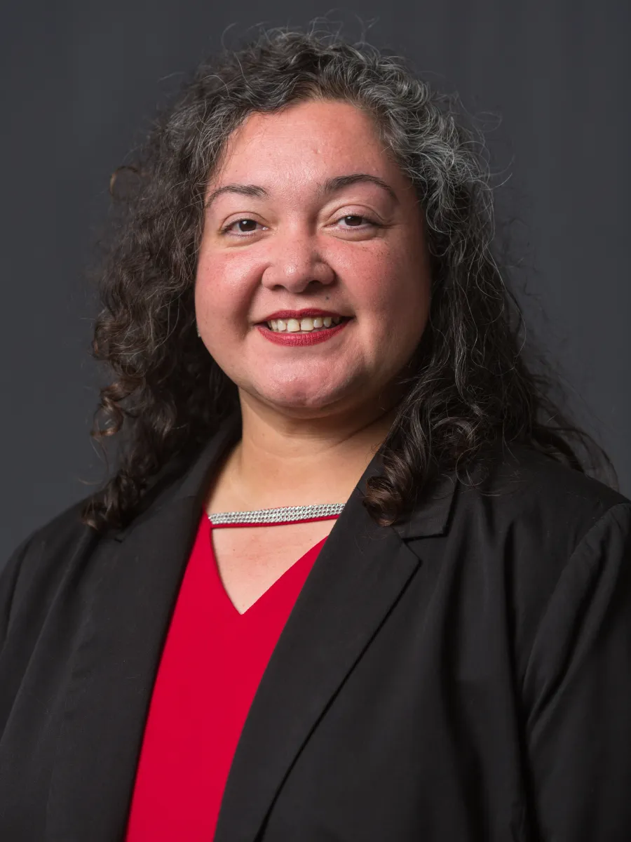 Marlina Martinez - Smiling woman with curly hair, wearing a black blazer over a red top, against a gray background.