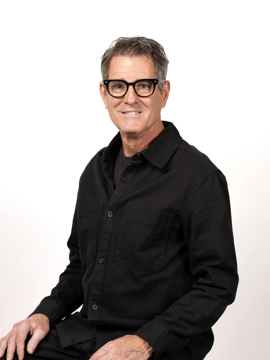 Michael Letts - Man with short hair and glasses, wearing a black shirt, sitting on a stool against a plain white background.