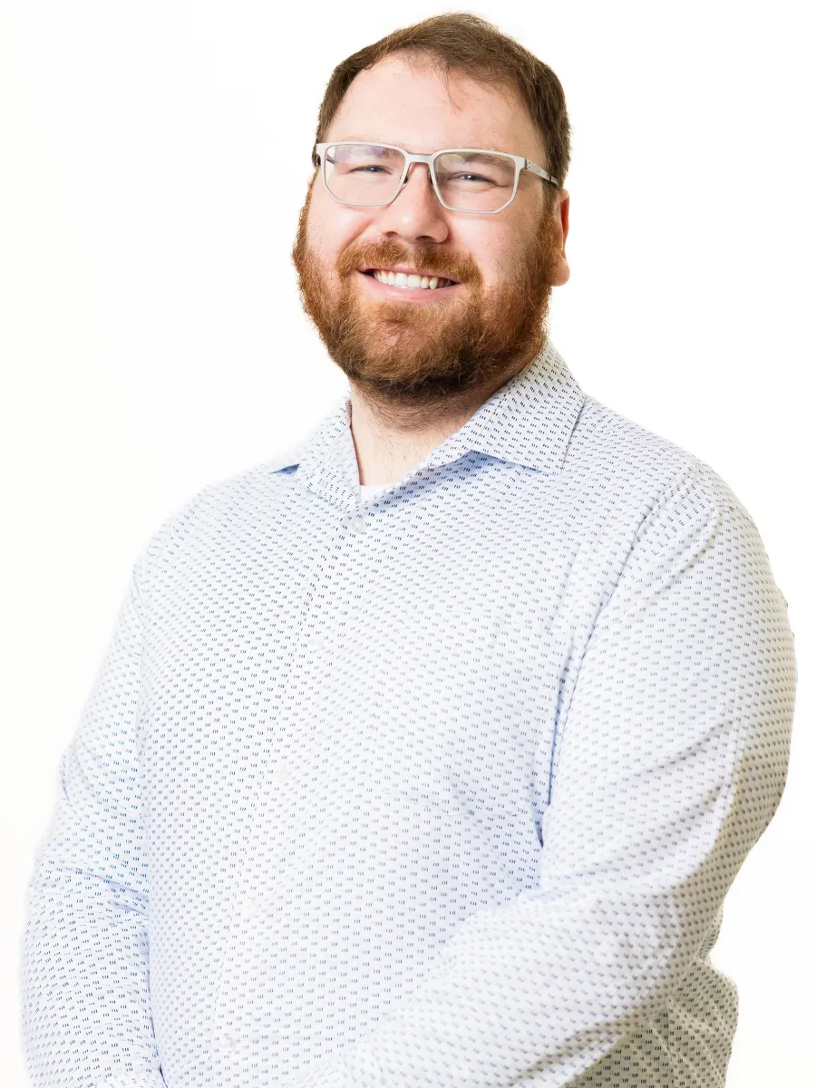 Simon Moesch - A smiling person with glasses and a beard stands against a plain white background. He is wearing a light-colored, patterned button-up shirt and has his hands clasped in front of them.