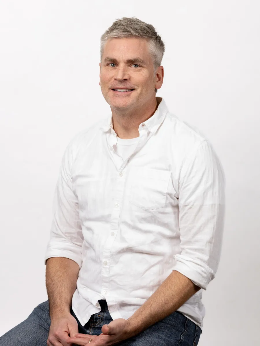 A man sitting against a plain white background, wearing a white shirt over a white T-shirt and blue jeans. He has short, gray hair and is smiling with hands resting on his lap.
