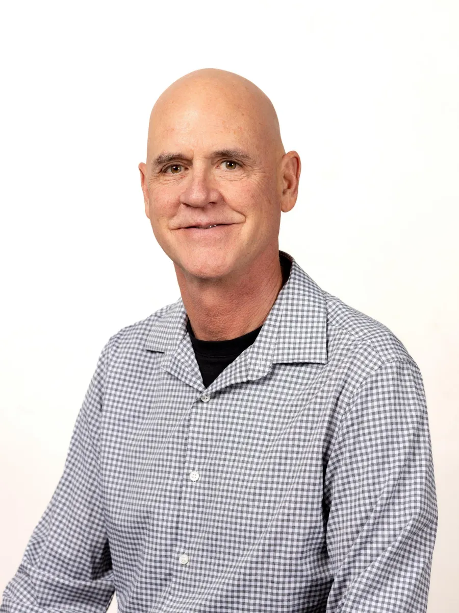 Steven Leuthold - A man with a slight smile is wearing a black shirt under a gray and white checkered button-up shirt. He is sitting against a plain white background.