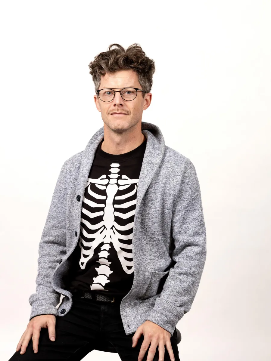Taimur Cleary -  A person with short, wavy hair and glasses is wearing a skeleton print shirt under a gray cardigan. They are sitting against a plain white background with a relaxed expression.