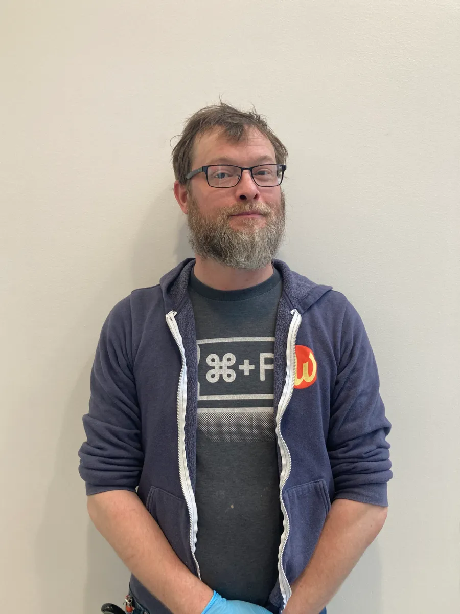 Chris Moore - A man with glasses and a beard stands against a plain light-colored wall. He wears a blue zip-up hoodie, a dark t-shirt with symbols, and blue gloves. He is looking at the camera with a slight smile.