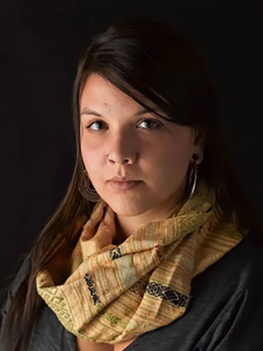 Christine Lenzen - A woman with long dark hair, wearing hoop earrings and a patterned yellow scarf, looks at the camera with a neutral expression against a dark background.