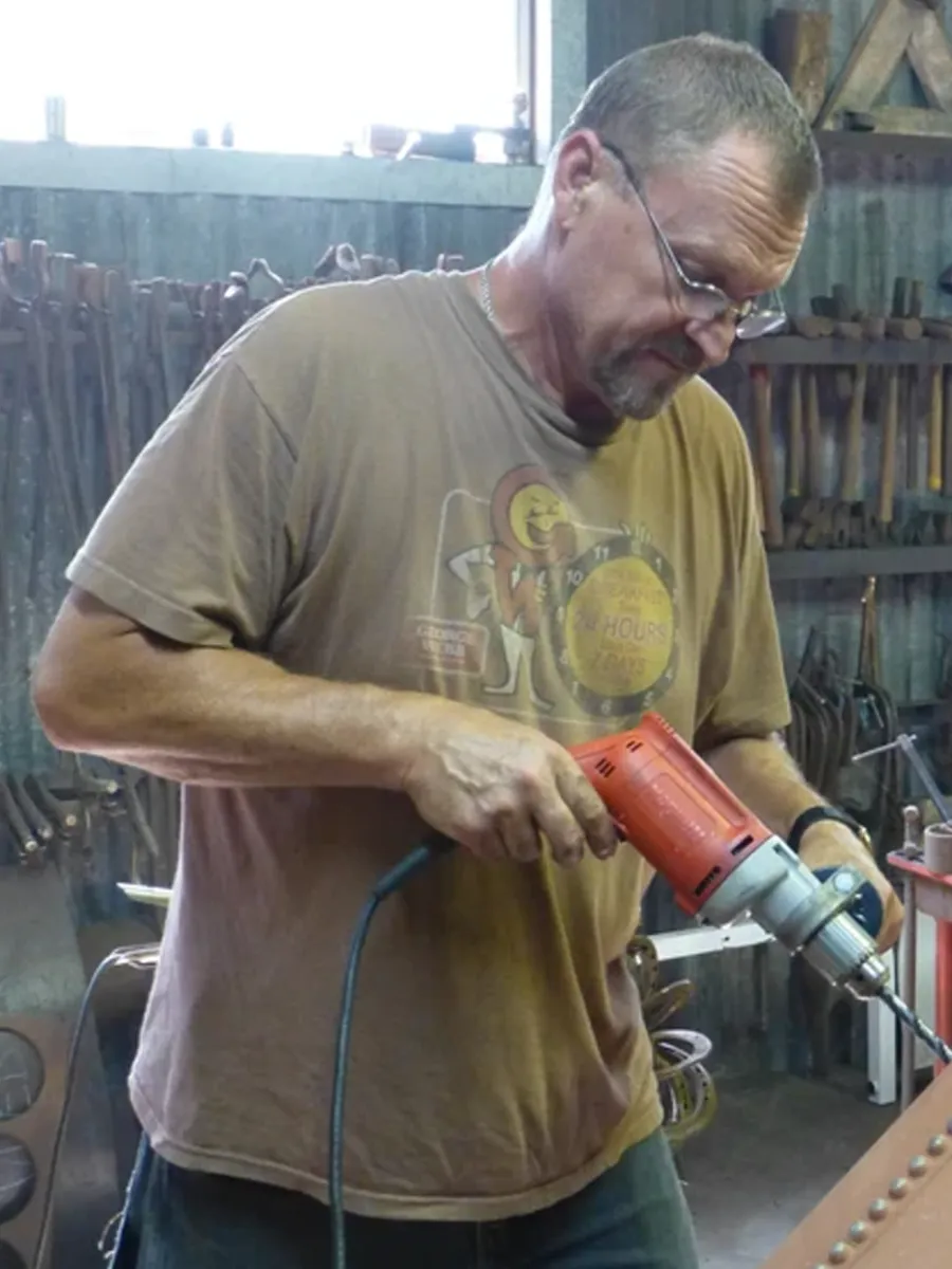 Dale Wedig - A man wearing glasses and a brown T-shirt uses a red power drill in a workshop filled with various tools hanging on the wall.