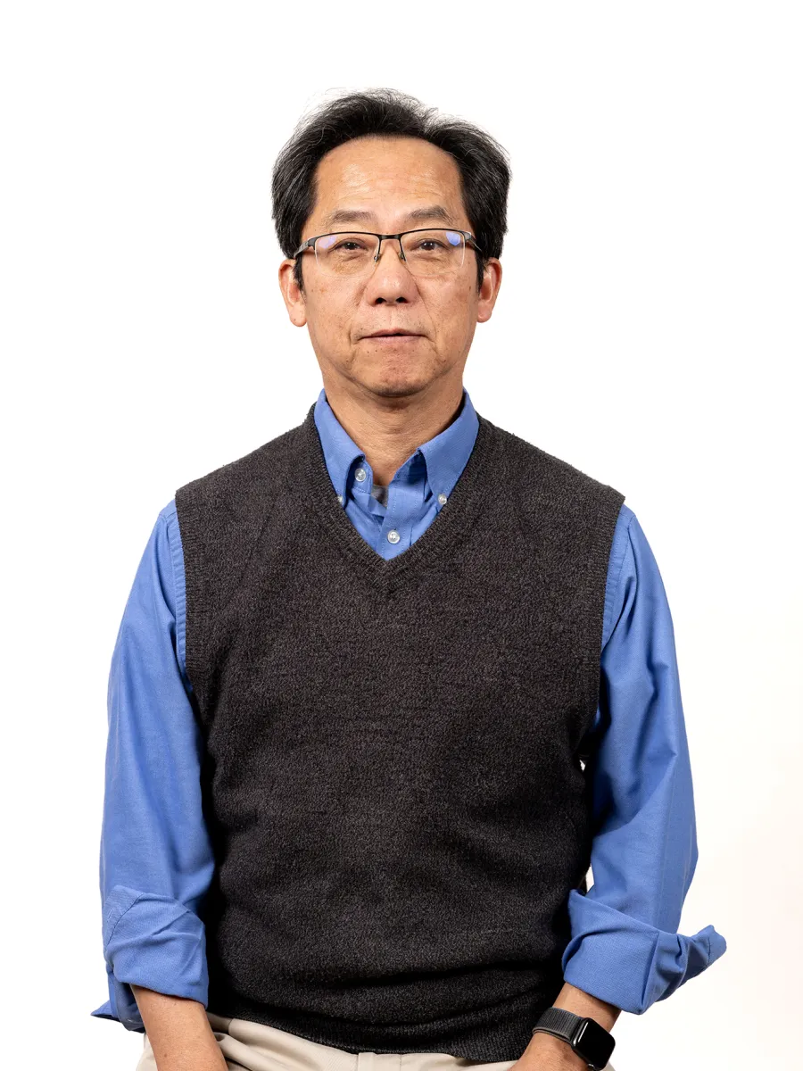 Dr. Mitsutoshi Oba - A man with short dark hair and glasses wearing a blue shirt and a dark gray sweater vest, standing against a plain white background.