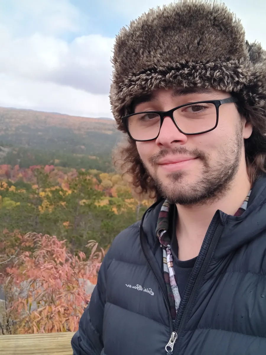 Harrison Stine - A person with glasses and a furry hat takes a selfie outdoors, wearing a black jacket. Behind them are autumn trees with colorful foliage and rolling hills under a partly cloudy sky.