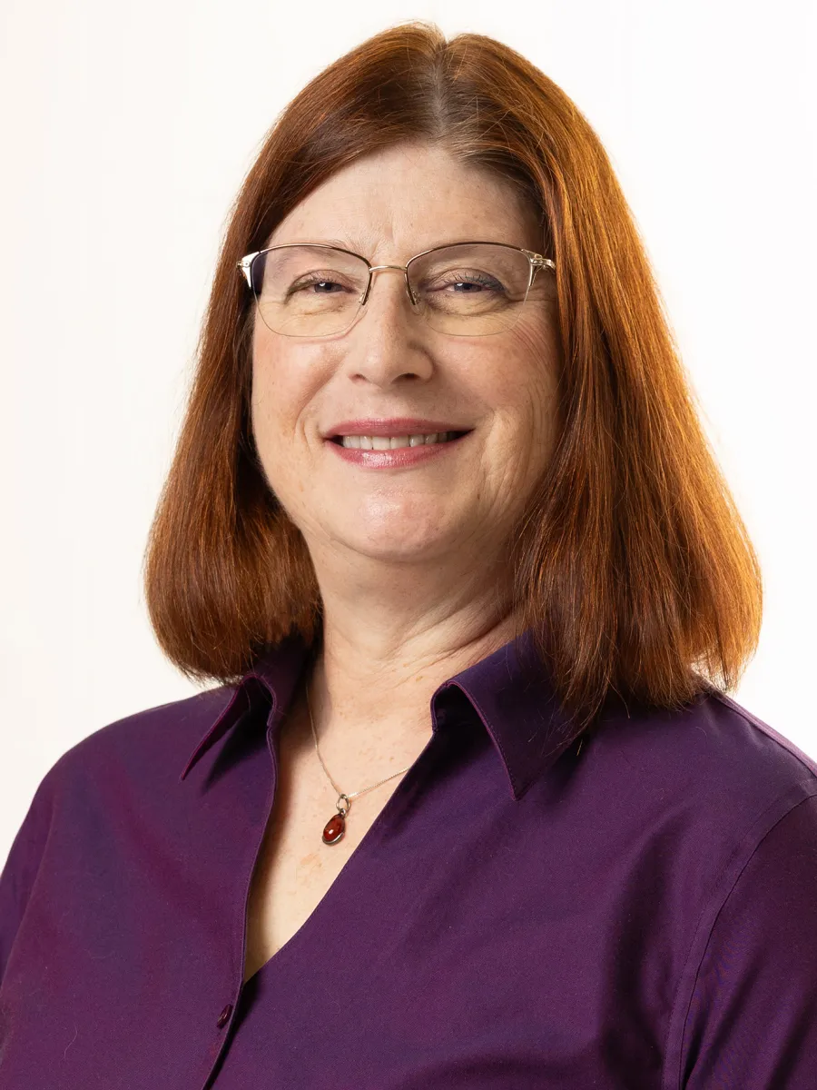 Jane Milkie - A woman with straight, shoulder-length auburn hair, wearing glasses, a purple collared shirt, and a pendant necklace, smiling at the camera against a plain white background.