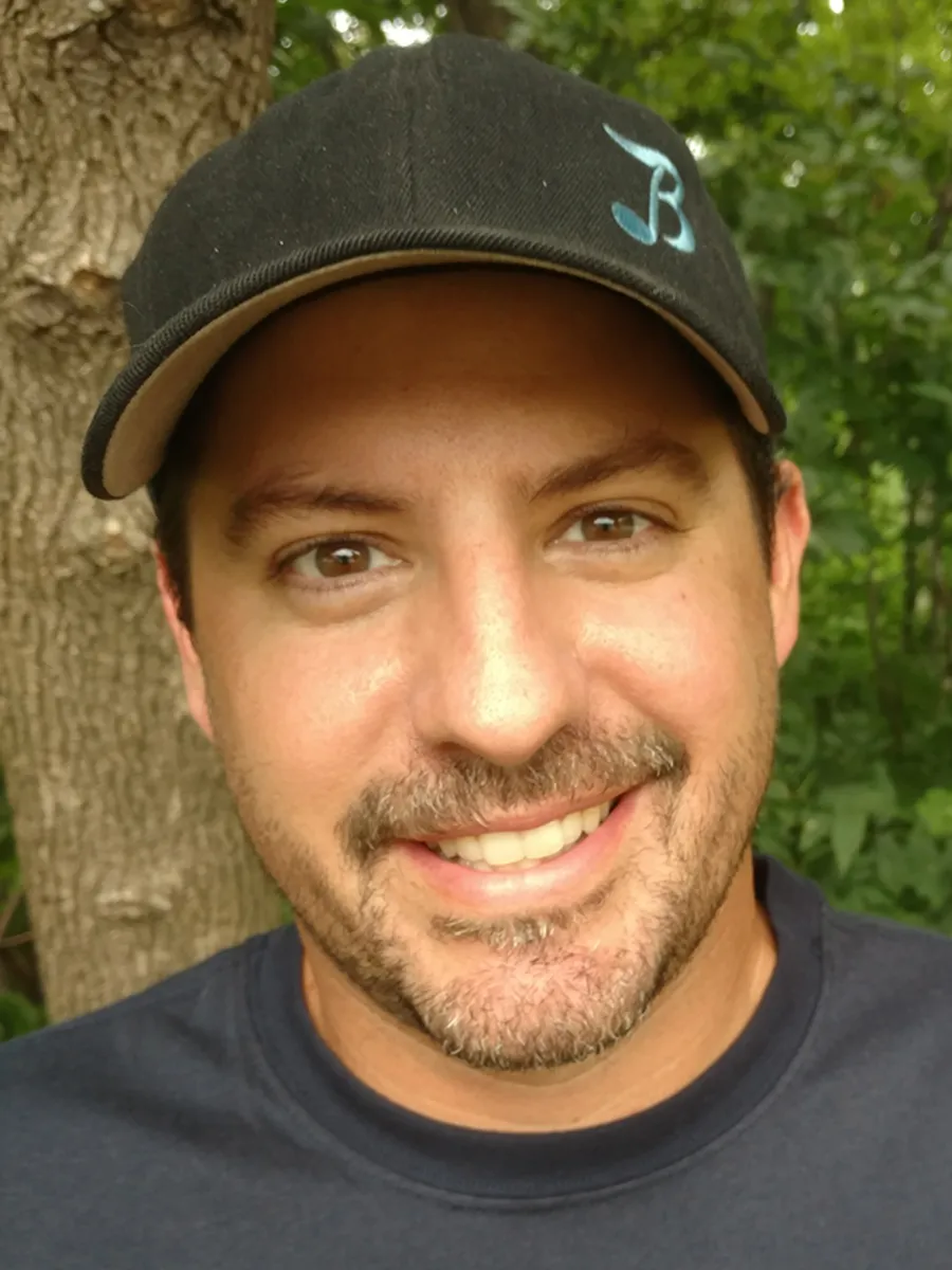 Jason Scneider - A man with brown eyes, and a short beard smiles outdoors near a tree. He wears a black baseball cap with a blue logo and a dark shirt. Green foliage is visible in the background.