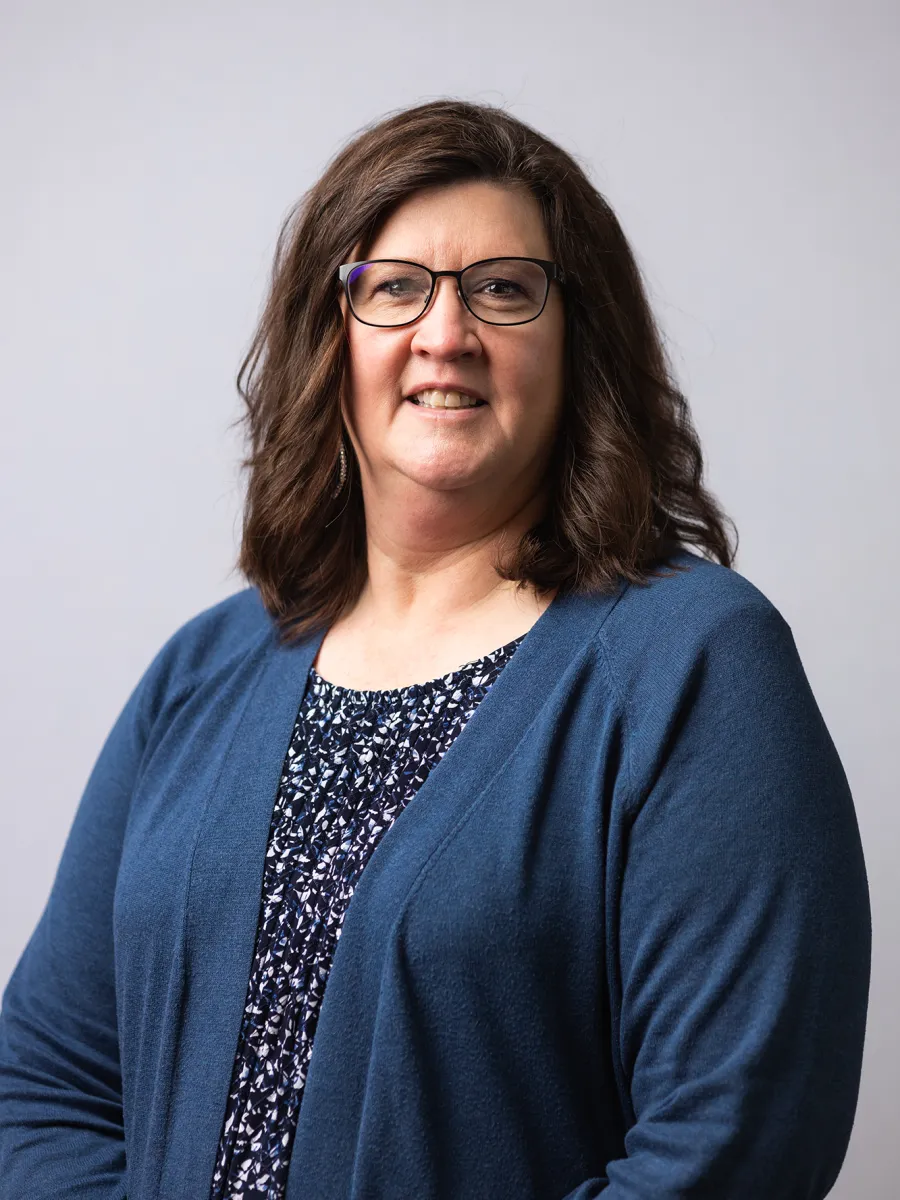 Karey LaJoye - A woman with shoulder-length brown hair, wearing glasses, a blue cardigan, and a patterned blouse, smiles at the camera against a plain light gray background.