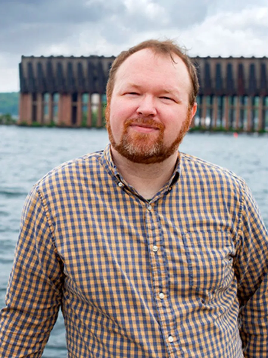 Keith Ellis - A man with short brown hair and a beard, wearing a yellow and blue plaid shirt, stands in front of a body of water with an industrial structure and cloudy sky in the background.