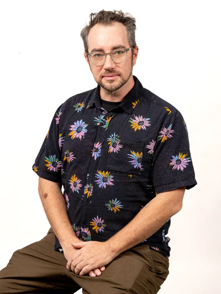 Nathan Bett - A person with short hair and glasses, wearing a black floral shirt and brown pants, sits on a stool against a plain white background, looking at the camera.