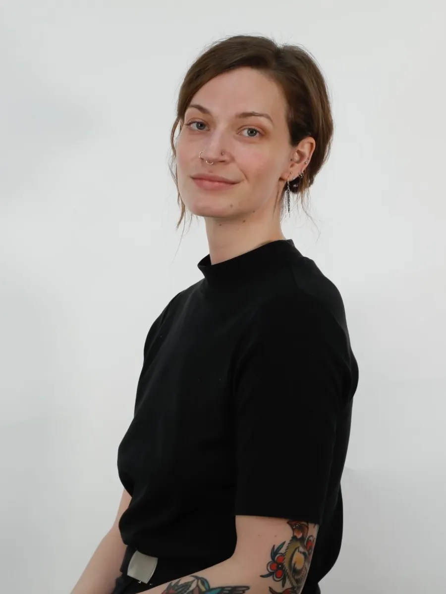 Sammie Hardewig - A woman with light skin and brown hair, wearing a black short-sleeved top, sits against a plain white background. She has tattoos on her left arm and a septum piercing, and is smiling softly at the camera.