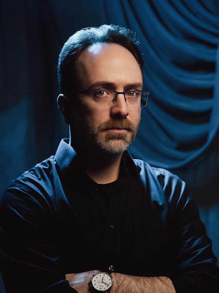 Steve Hughes - A man with short dark hair, glasses, and a beard is wearing a black shirt and a wristwatch. He stands with arms crossed against a blue backdrop with draped fabric, looking seriously at the camera.