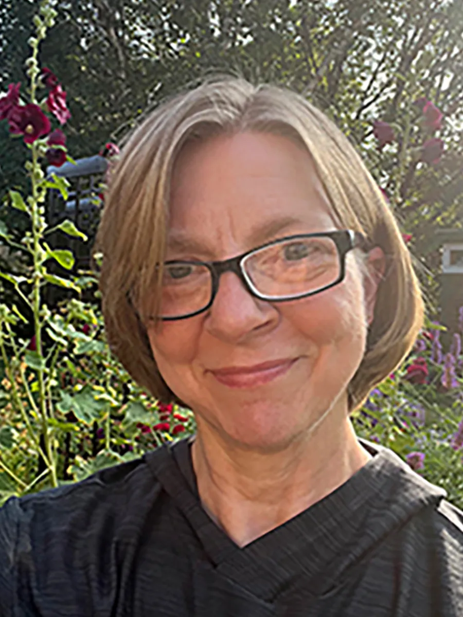 Tracy Wascom - A woman with short, light brown hair and glasses smiles at the camera while standing outdoors in a garden with purple and red flowers and greenery in the background.