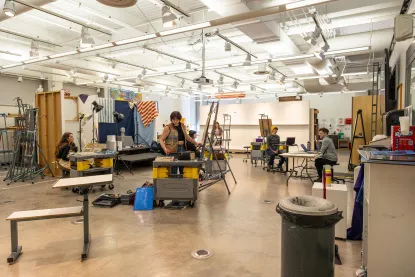 A spacious art studio with five people working. Some are at easels and others are on computers. Art supplies and equipment are scattered around. The walls have art pieces, and the ceiling has track lighting.