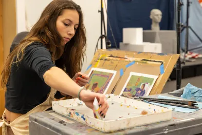 Woman with long hair painting in a studio. She is focused on a paint palette in front of her, surrounded by various art tools. Behind her are artworks and a bust on a stand. The scene is well-lit with studio lights.