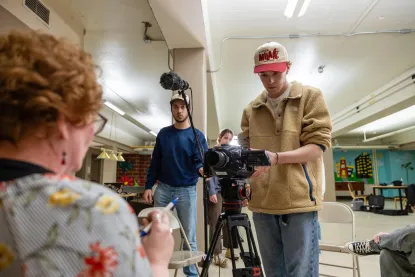 A person wearing a red and white hat operates a camera on a tripod in a room with bright lighting. Another person stands nearby. A seated person in the foreground faces away, holding a pen and paper. A microphone is positioned above them.