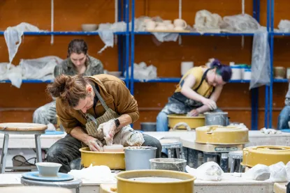 Three people are focused on shaping pottery on pottery wheels in a studio. The room has shelves with pottery and supplies in the background.