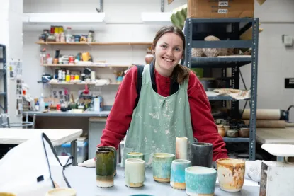 A person in a pottery studio smiles at the camera. They are wearing a red shirt and a green apron, standing behind a table with various colored ceramic cups. Shelves in the background hold pottery materials and tools.
