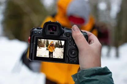A hand holds a camera, capturing a person in a yellow jacket standing in a snowy landscape. The camera's screen displays the scene, focusing on the person holding ski poles. The background is blurred, with trees visible.