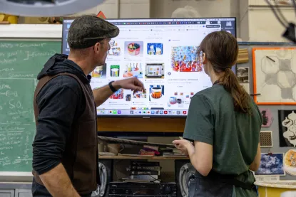 A man and a woman stand in front of a large monitor displaying images of colorful art supplies. The man points at the screen while they discuss. A chalkboard with notes and shelves filled with tools are in the background.