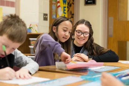 A young girl points at a notebook while a woman beside her smiles. They are sitting at a classroom table with other children visible.