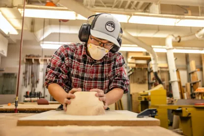A person in protective gear, including earmuffs, a cap, and a face mask, is sanding a wooden piece in a workshop. They're wearing a red plaid shirt, and various tools and equipment are visible in the background.