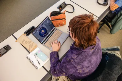 Person with short auburn hair and glasses working on a laptop at a desk, designing a circular graphic. Nearby are a notebook, phone, and an orange bag. Overhead view.