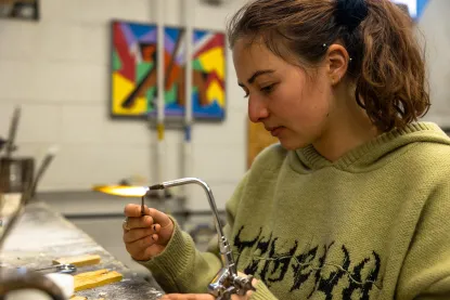 A person wearing a green sweater uses a handheld blowtorch in a workshop. They focus intently on their work. Colorful abstract paintings are visible on the wall in the background.