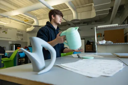 A person in a classroom holds a light blue sculptural object resembling a pitcher. Nearby are sketches and another similar object on the table.