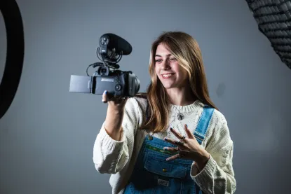 A young woman wearing a white sweater and denim overalls smiles while holding a video camera with a microphone attached, standing in a studio with professional lighting equipment.