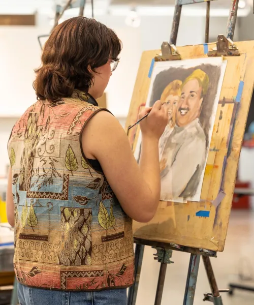 A person with short brown hair stands in front of an easel, painting a portrait of two smiling people. They wear a colorful, patterned vest and have star tattoos on their arm. The room has art supplies and storage cabinets in the background.