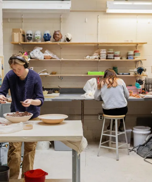 Two people work in a pottery studio. One person shapes clay on a table in the foreground, while the other person sits at a workstation in the background. Shelves with pottery and tools surround them. The room is brightly lit.