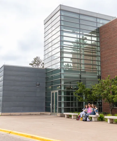A modern building with glass and brick walls, featuring a group of people sitting on a bench near the entrance. The sky is cloudy, and there is a pathway and greenery around the structure.