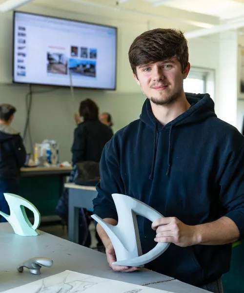 A young man in a black hoodie stands at a table holding a curved sculptural object in a classroom. Behind him, people converse and a large screen displays images and text. Sketches and similar objects are on the table.